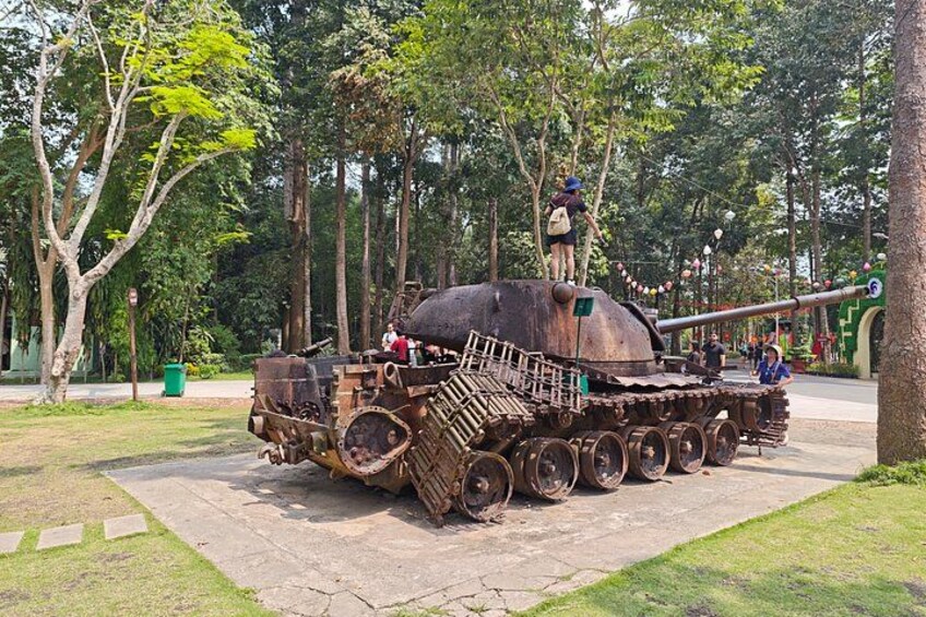 A large, rusted military tank on display in a park with trees and a paved walkway. 2