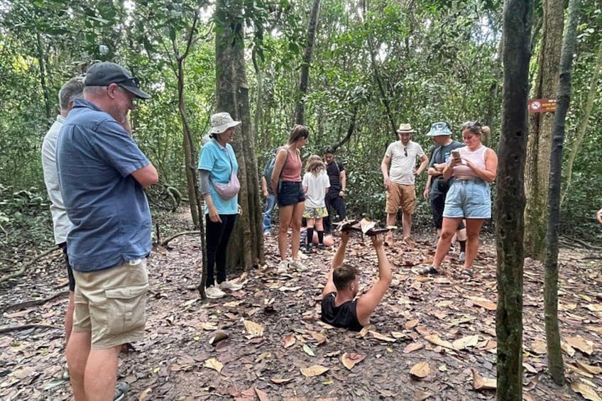 Un grupo de personas en un área boscosa, algunas de pie y otras sentadas en el suelo, participando en una actividad al aire libre. 3
