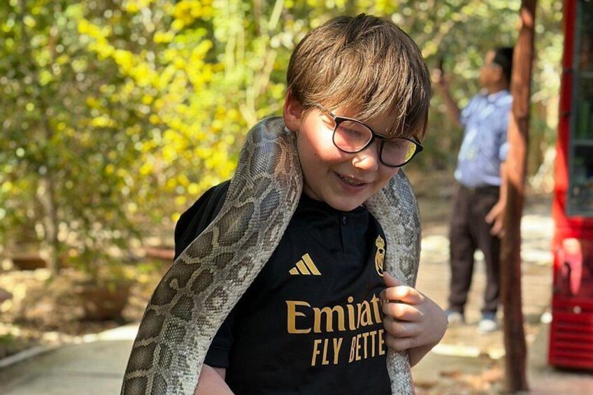Eine Person mit schwarzem Hemd, auf dem 'Emirates' und 'Fly Better' steht, hält eine Schlange. 2