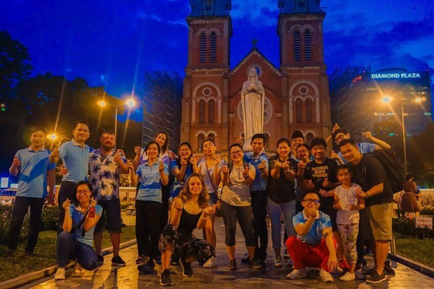 Un gruppo di persone che posa davanti a una chiesa con una grande statua, illuminata dalla luce serale. 1