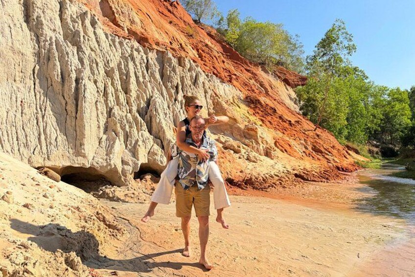 Una persona porta un'altra sulla schiena, camminando su un sentiero sabbioso accanto a un fiume con una scogliera rocciosa sullo sfondo. 2