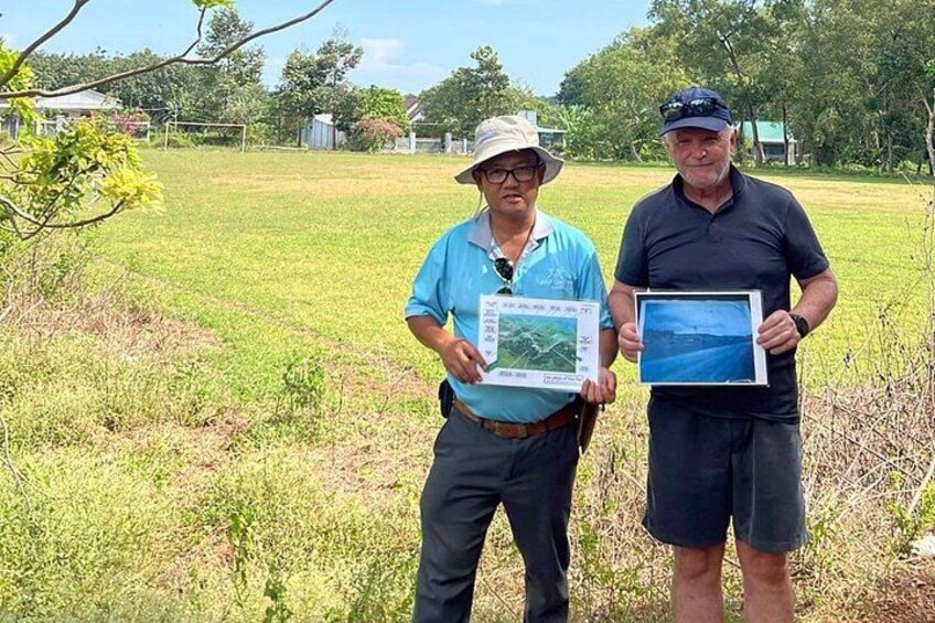 Two men holding framed photographs in a grassy field with trees and houses in the background. 5