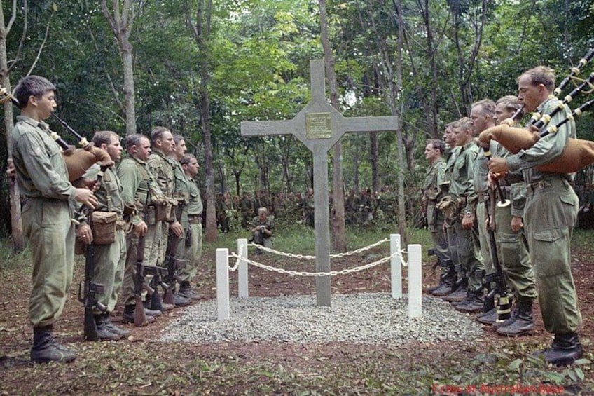 Soldiers in uniform standing in a line with rifles, a cross-shaped monument in the center, and a chain-link fence in a wooded area. 1