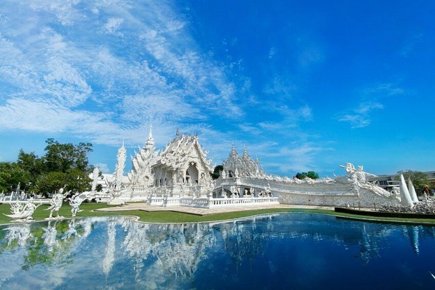 A white temple complex with intricate architecture, a large reflective water body in the foreground, and a clear blue sky. 3