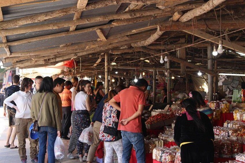Un mercado con vigas de madera y luces colgantes, gente comprando y diversos productos a la vista. 5