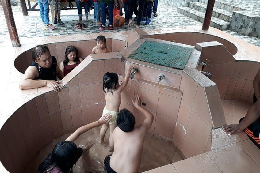 Un groupe de personnes se trouve dans un espace de bains public, dont les murs sont recouverts de carreaux et qui est agrémenté d’une fontaine centrale. 1