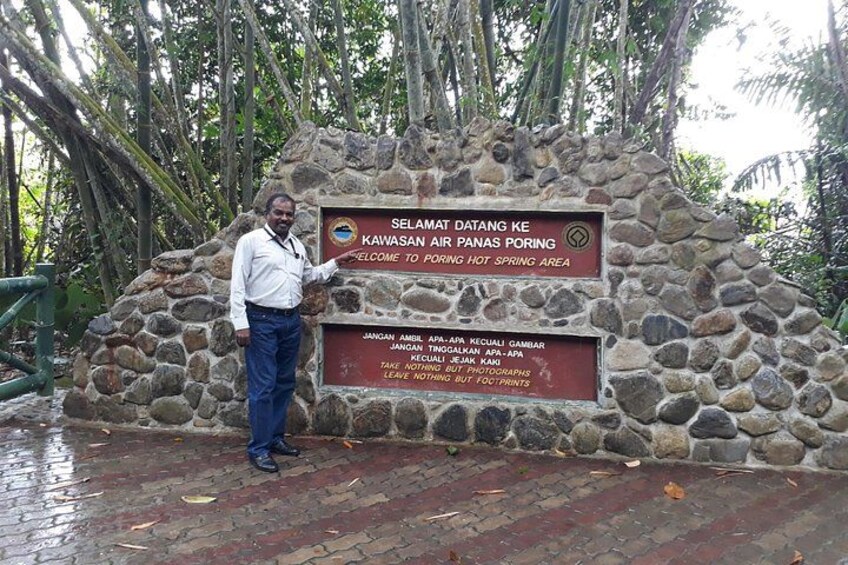 Un homme se tient devant un mur de pierre sur lequel est affiché un panneau indiquant : « Selamat Datang ke Kawasan Air Panas Poring » et qui précise les heures d’ouverture de la zone des sources chaudes. 4