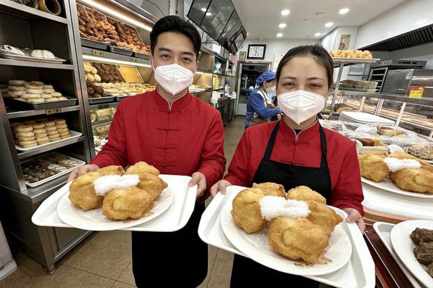 Two individuals in red uniforms holding trays of fried dough pastries in a bakery. 2