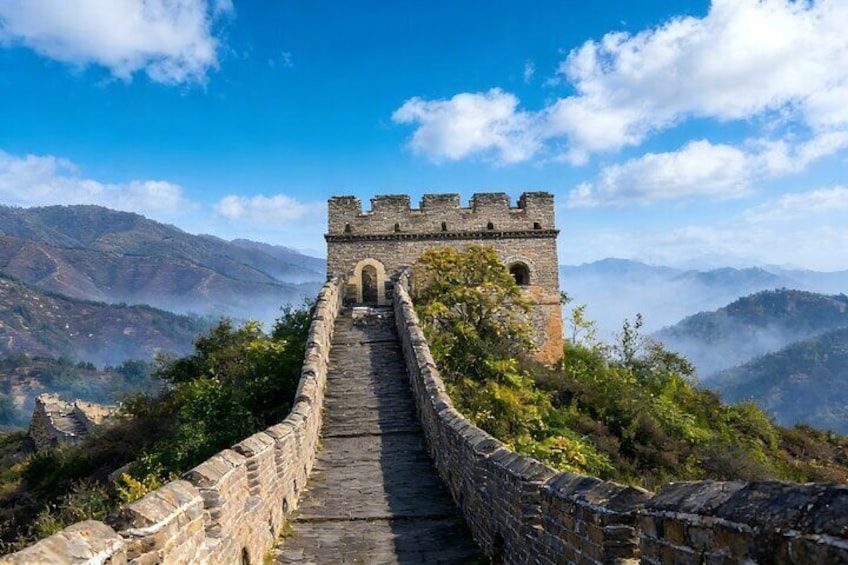 A stone wall leading to a castle-like structure with a clear sky and mountains in the background. 1