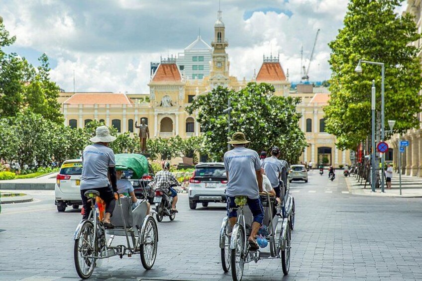 Una strada con persone che vanno in bicicletta e in moto, un edificio con il tetto rosso e alberi che fiancheggiano la strada. 5