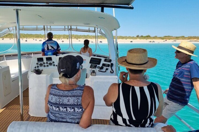 Ein Boot mit Passagieren genießt einen sonnigen Tag, ein Strand mit Personen im Hintergrund und klares, blaues Wasser. 3