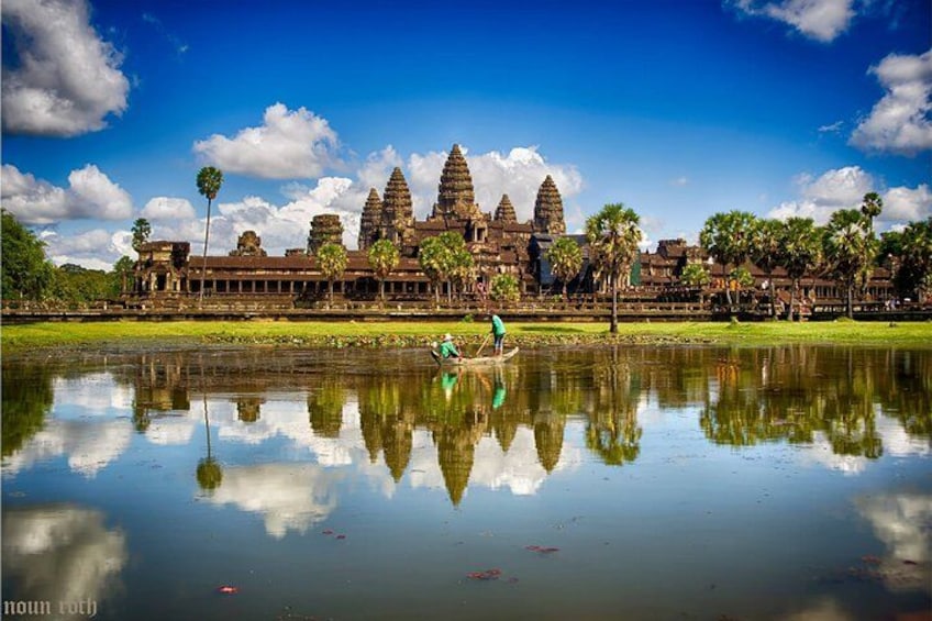 Angkor Wat temple, a large historical structure, with a reflection in the water and a person paddling a boat. 1