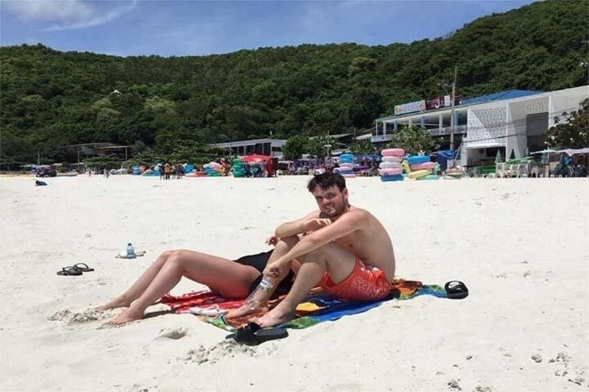 Eine Person sitzt auf einem Strandtuch, im Hintergrund Strand und Gebäude. 4