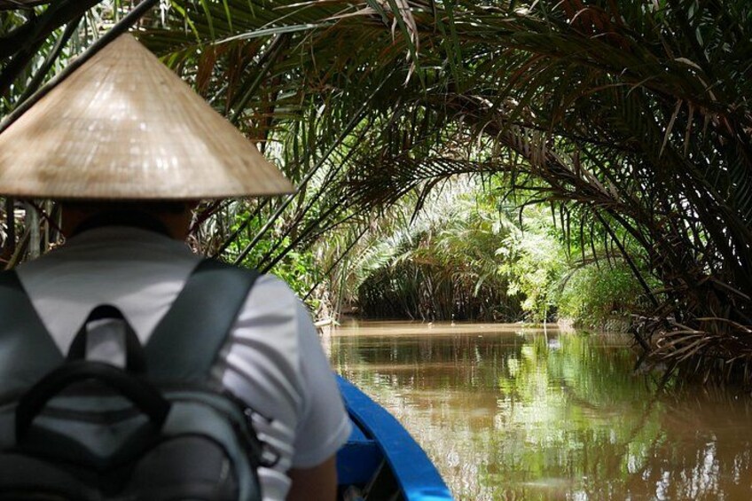 A person in a conical hat paddling a blue boat through a waterway surrounded by dense vegetation. 2