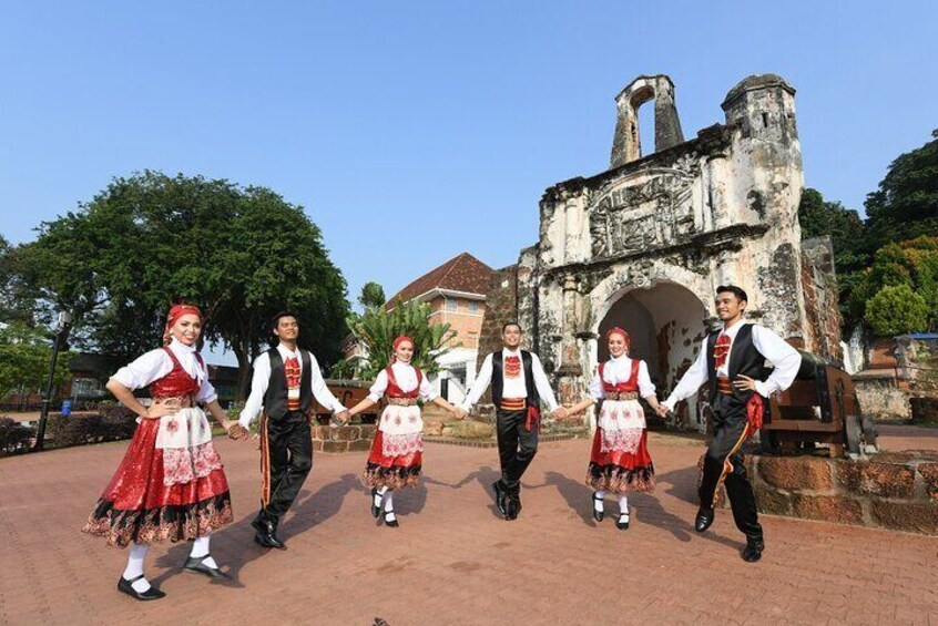 Five people in traditional attire performing a dance in front of a historical stone structure. 1