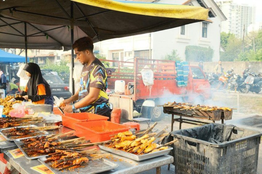 Un vendedor de comida callejera asando brochetas bajo una tolda, con una camioneta roja y motocicletas al fondo. 5