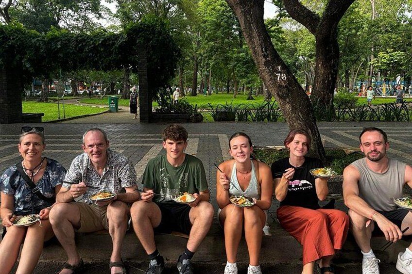 A group of people sitting on a bench in a park with trees and grass in the background. 2