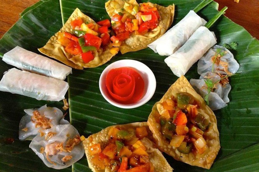 A variety of spring rolls arranged on a banana leaf with a red rose-shaped garnish. 3
