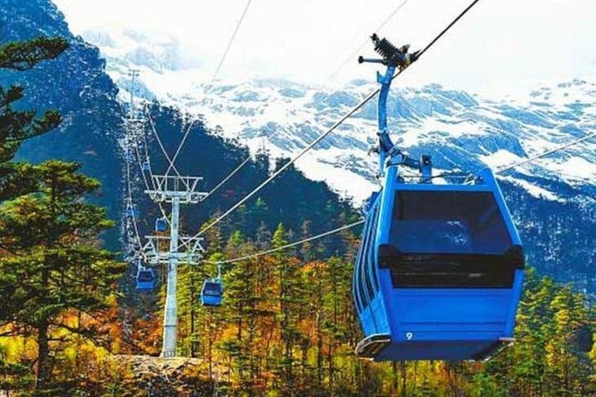 Una funivia sospesa sopra una zona montuosa ricoperta di boschi, con cime innevate sullo sfondo. 2