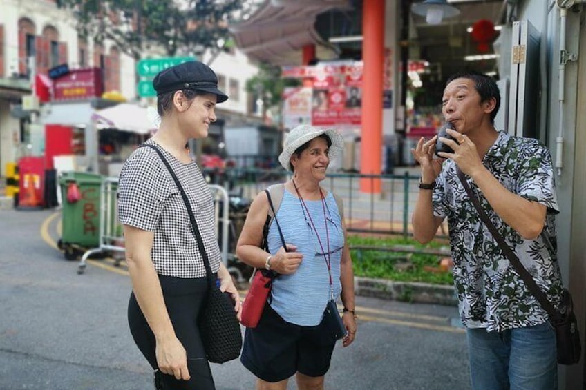 Three people walking on a street with shops and a red pillar in the background. 1