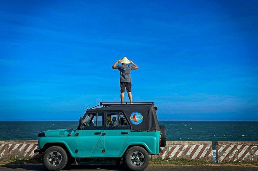 A turquoise off-road vehicle with a person standing on top, a clear blue sky, and a calm sea in the background. 1