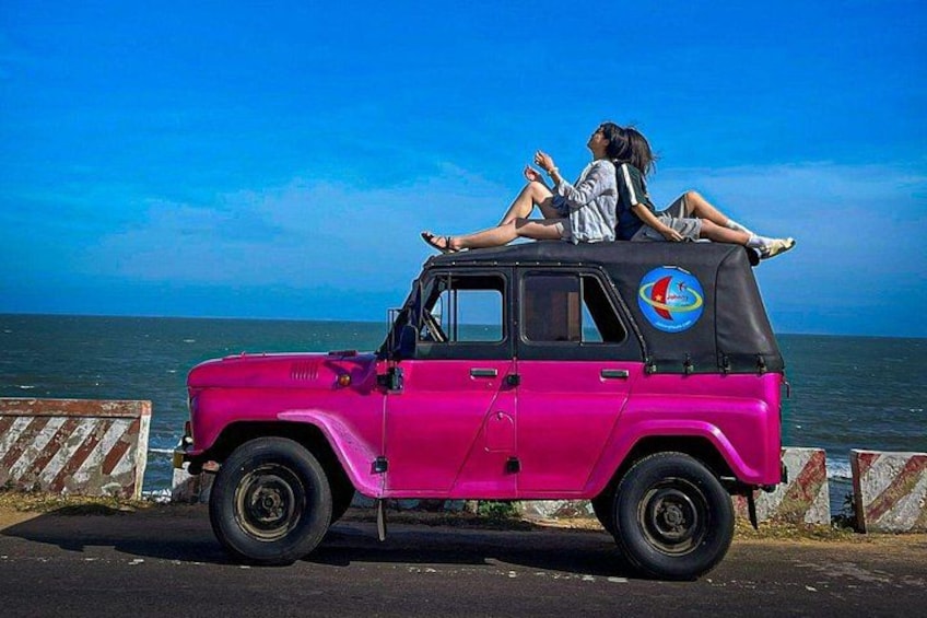 A pink jeep parked by the sea with two people sitting on the roof. 2