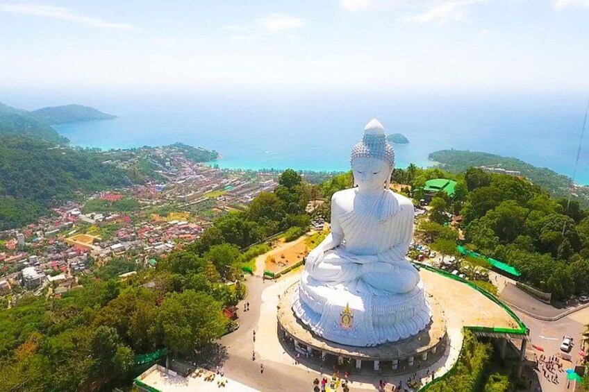 A large white Buddha statue surrounded by a circular walkway, with a coastal town and mountains in the background. 1