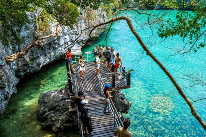 A wooden platform with people standing and walking on it, overlooking a clear blue body of water surrounded by rocky cliffs. 4