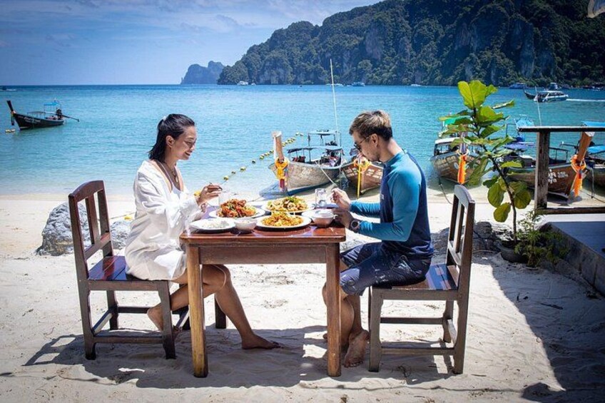 Deux personnes sont assises à une table en bois sur une plage, avec des bateaux en arrière-plan. 1