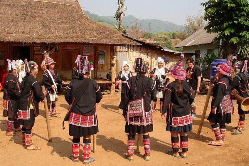 A group of people in traditional attire performing a dance in front of a rustic thatched-roof building. 3