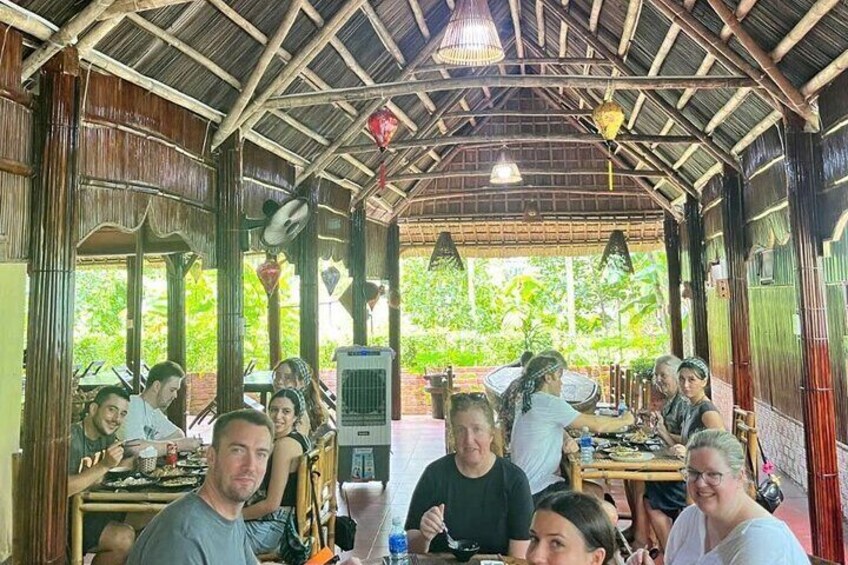 Un groupe de personnes est réuni autour d’une table à l’intérieur d’une structure couverte, dotée d’un toit de chaume et de poutres en bois. 2