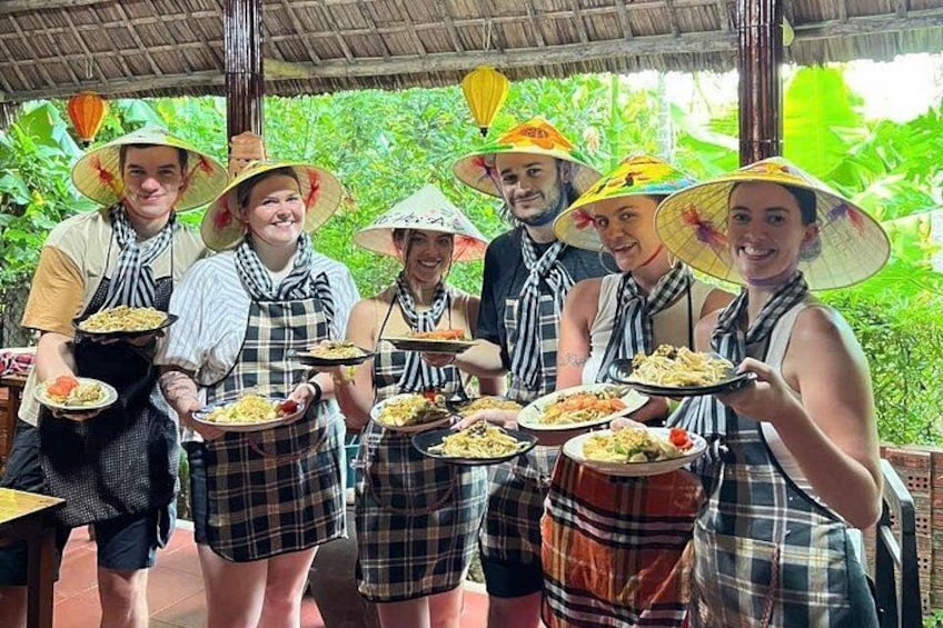 Een groep mensen in traditionele kleding die schalen met eten vasthouden, staand onder een rieten dak. 1