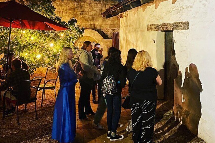 People gathered outside a building with a red umbrella, a stone wall, and a tree with lights. 2