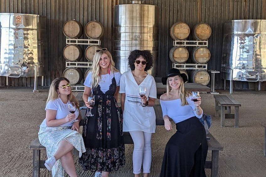 Eine Gruppe von Menschen sitzt und steht draußen, Wein gläser in der Hand, mit Fässern und einer großen Metallkonstruktion im Hintergrund. 2