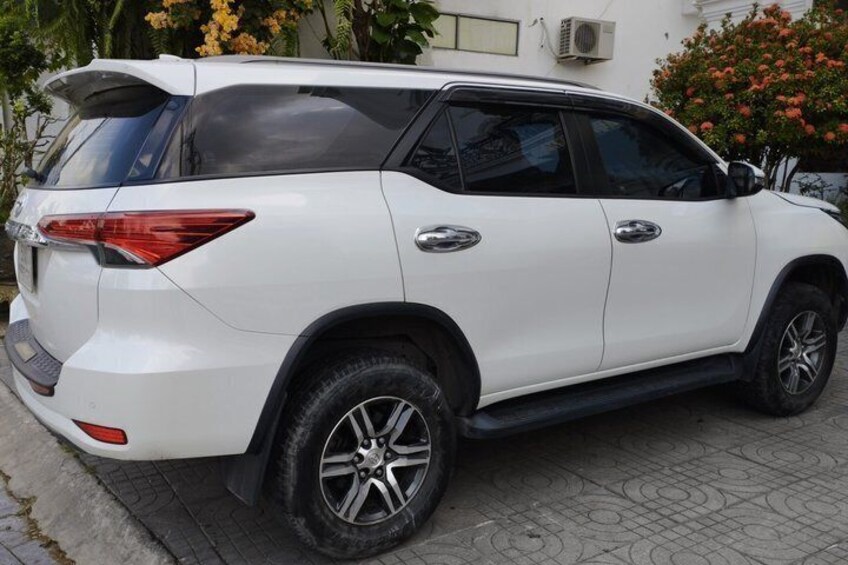 A white SUV parked on a paved surface with a building and flowering plants in the background. 2