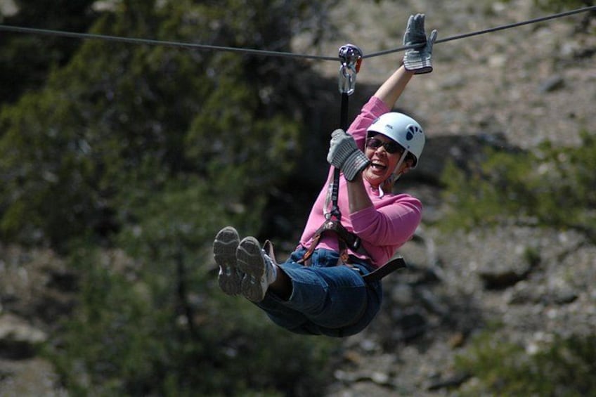 En person suser ned af en zipline, iført hjelm og handsker, med et klippefyldt landskab i baggrunden. 1