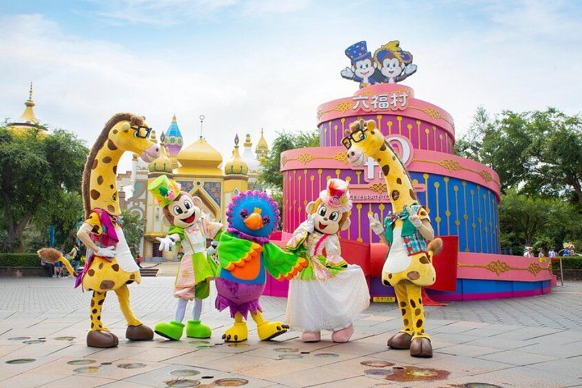 A giraffe mascot, a clown, and a character in a green dress standing in front of a colorful building with a sign that reads 'Happy Birthday'. 3