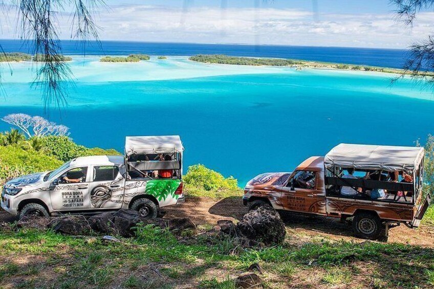 Deux véhicules de safari sont stationnés sur un chemin de terre, avec une mer turquoise et une végétation luxuriante en arrière-plan. 3