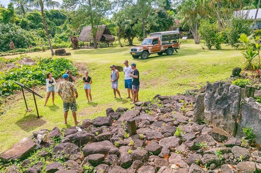 Un groupe de personnes se tient sur une zone herbeuse, avec un terrain rocheux au premier plan, une construction à toit de chaume et un véhicule à l’arrière-plan. 2