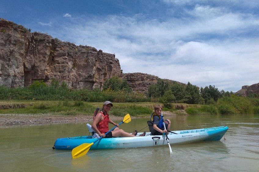 Two people kayaking on a river with rocky cliffs in the background. 2