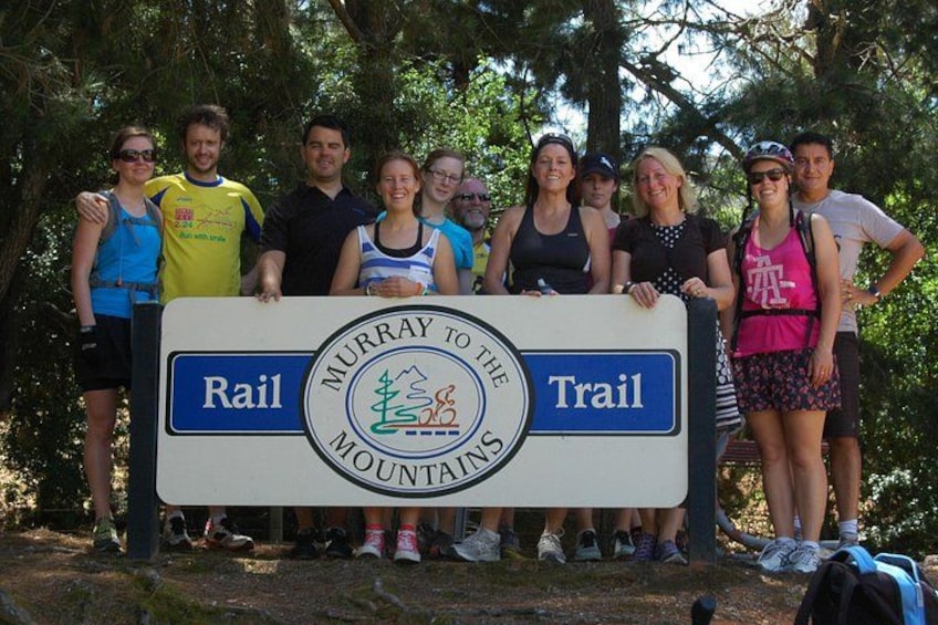 A group of people standing in a forested area with a sign that reads 'Murray to the Mountains Trail.' 1