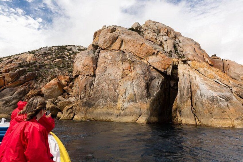 A rocky cliff with a cave entrance, a person in a red jacket, and a boat in the water. 3