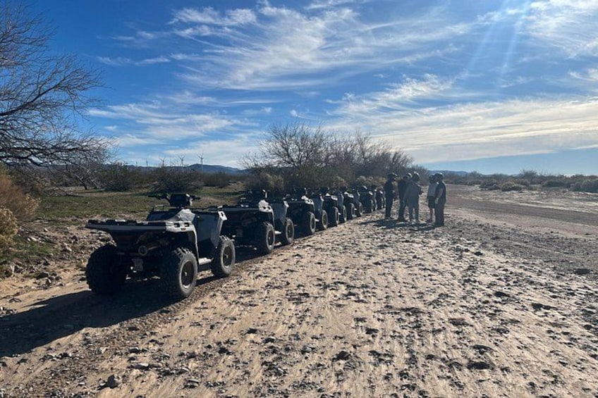 A line of ATVs parked on a dirt road with a group of people standing nearby. 2