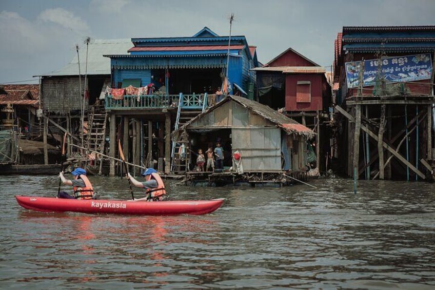 A red kayak with two paddlers on a calm river, surrounded by traditional wooden houses with colorful roofs. 2
