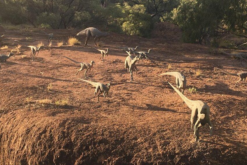 A group of dinosaurs in a dry, open landscape with sparse vegetation and trees in the background. 4