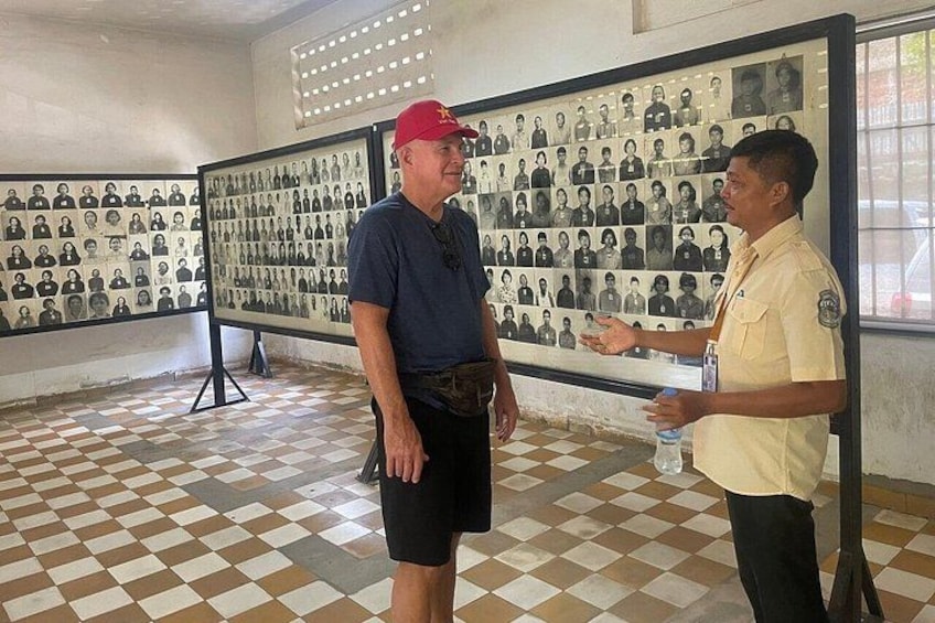 Due uomini in piedi in una stanza con pavimento piastrellato e un muro che espone numerose fotografie in bianco e nero. 3