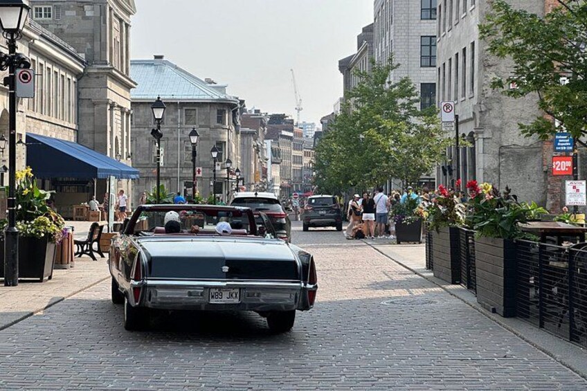 Ein Oldtimer-Cabriolet fährt auf einer Kopfsteinpflasterstraße entlang, gesäumt von Gebäuden und Fußgängern. 2