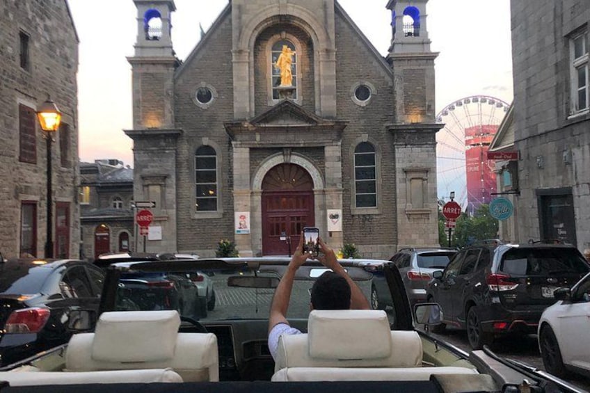 Eine Kirche mit einem großen Rundbogenportal und einem Glockenturm, umgeben von parkenden Autos und einem Riesenrad im Hintergrund. 5