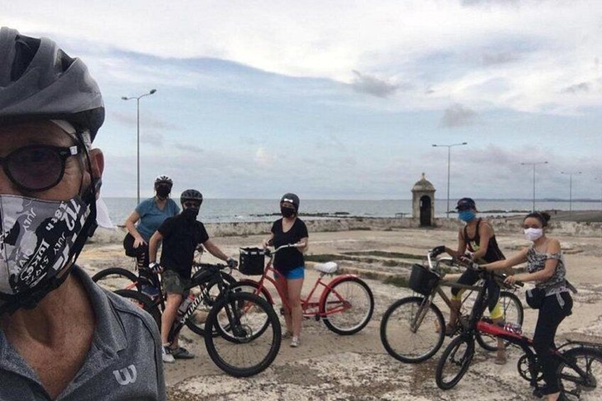A group of cyclists on a coastal path with a small structure in the background. 1