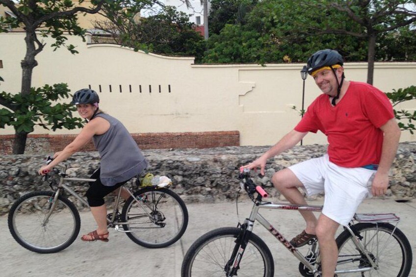 Two people riding bicycles on a paved path with a stone wall and trees in the background. 3